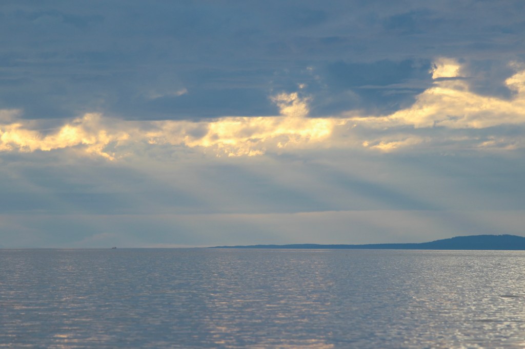 A tranquil seascape at sunset, featuring calm waters reflecting the sky with scattered clouds and rays of light breaking through, hinting at distant land on the horizon.