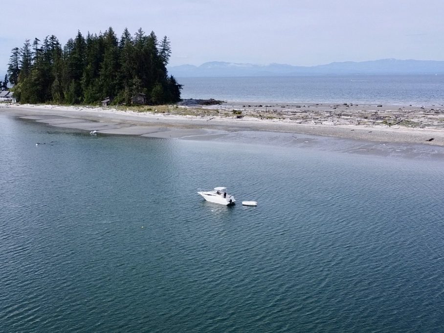 A scenic view of a calm bay with a small boat anchored in shallow water, surrounded by sandy beaches and a forested island in the background.